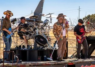 All four members of Wicked Truth on stage at an outdoor festival playing either the drums, guitar, bass or singing