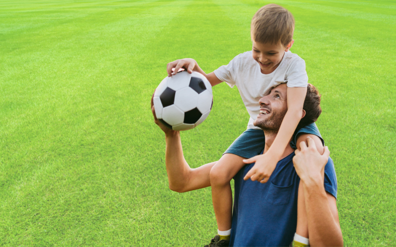 Man holding soccer ball with boy sitting on his shoulders